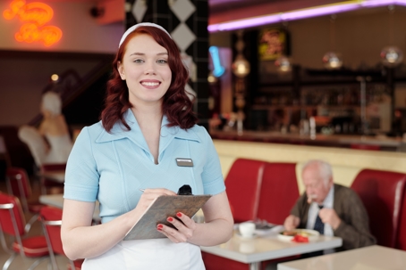 A cheerful waitress smiles at the camera while holding a notepad in a retro diner.